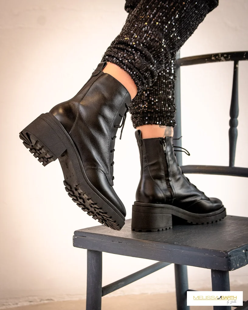Imagem das pernas de uma modelo usando a roupa preta com brilhos das imagens anteriores em pé sobre uma cadeira cor chumbo calçando uma bota tipo coturno preta com sola tratorada grossa e ziper na lateram com cadarço na parte superior. A bota é feita de couro e com pouco brilho.