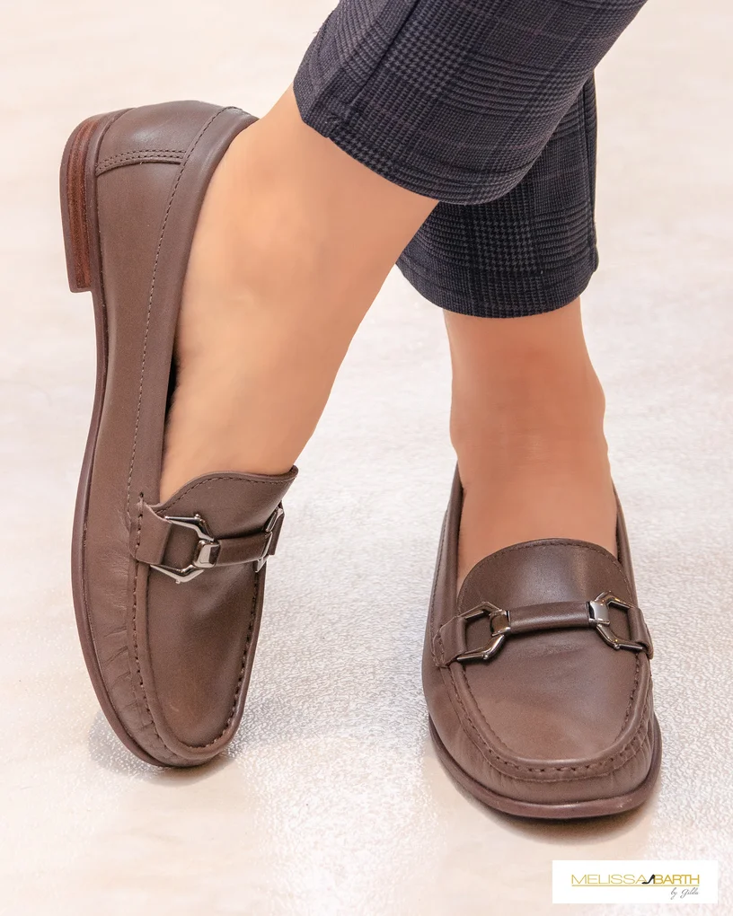 Foto enquadrada nos pés de uma modelo calçando um sapato mocassim cor marrom, um nos pés esta no chão e o outro esta na pontinha do pé.