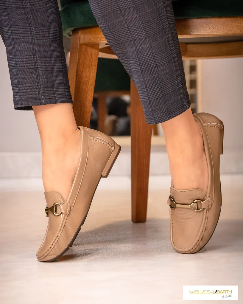 Imagem das pernas de uma modelo calçando um sapato mocassim cor bege, a modelo está sentada em uma cadeira e s sapatos estão inclinados na pontinha do pé.